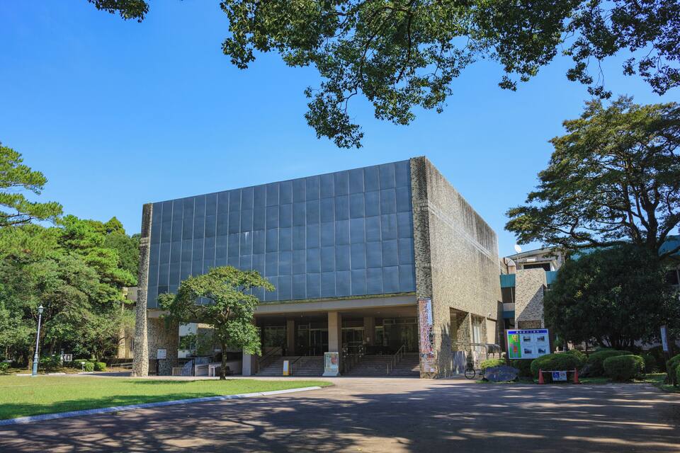 A modern building with a glass facade, surrounded by trees and a clear blue sky.