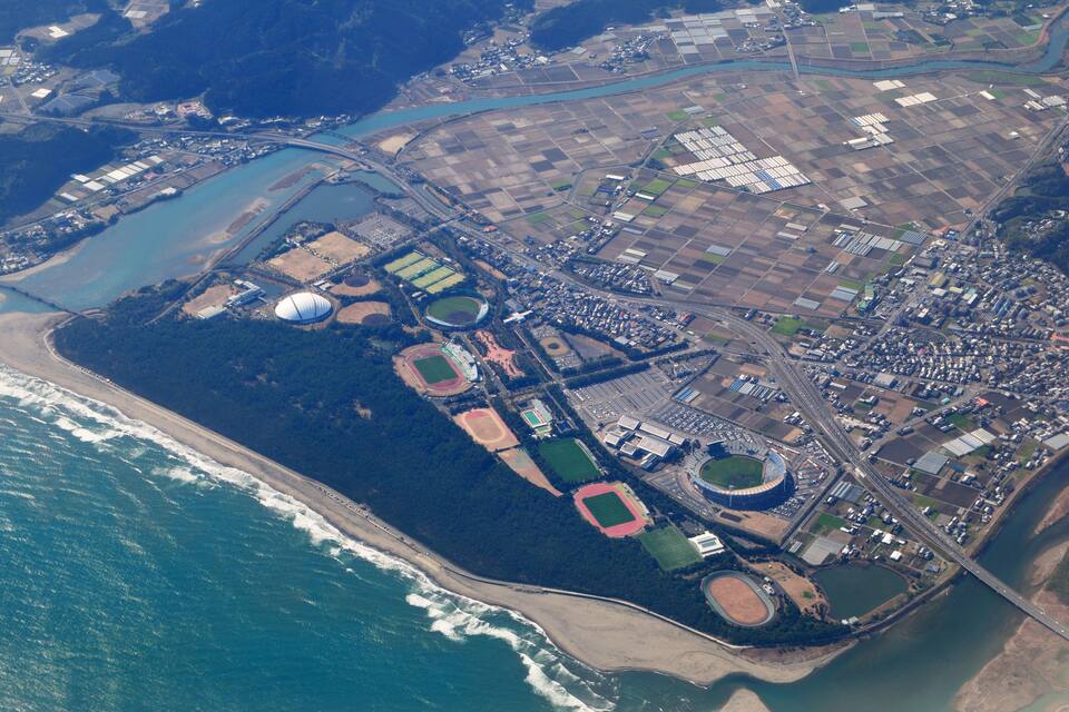 An aerial view of a coastal city with a stadium, sports fields, industrial areas, and a river.