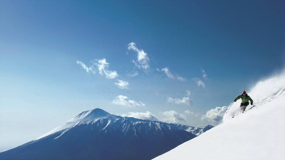 A skier on a snowy mountain slope with a clear sky and a snow-covered peak in the background.