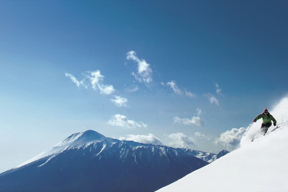 A skier on a snowy mountain slope with a clear sky and a snow-covered peak in the background.