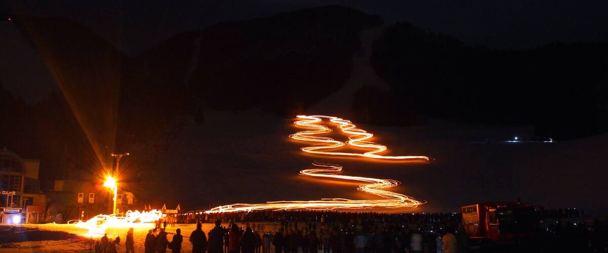 A night scene with a light trail forming a spiral pattern on the ground, a crowd of people, and a building in the background.