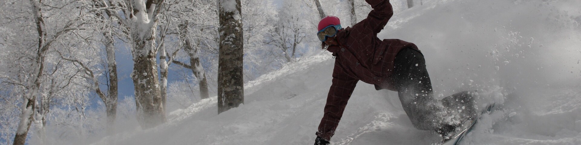 Un snowboarder con chaqueta roja y casco rosa en una pendiente nevada, con árboles al fondo.