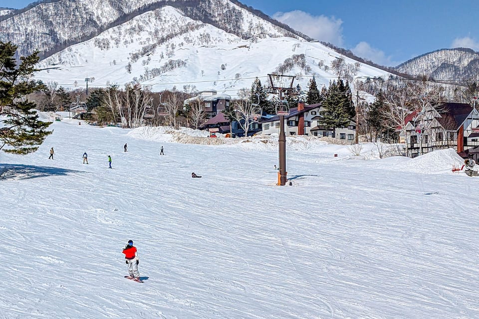 A snowy ski slope with skiers and a ski lift, surrounded by mountains and buildings.