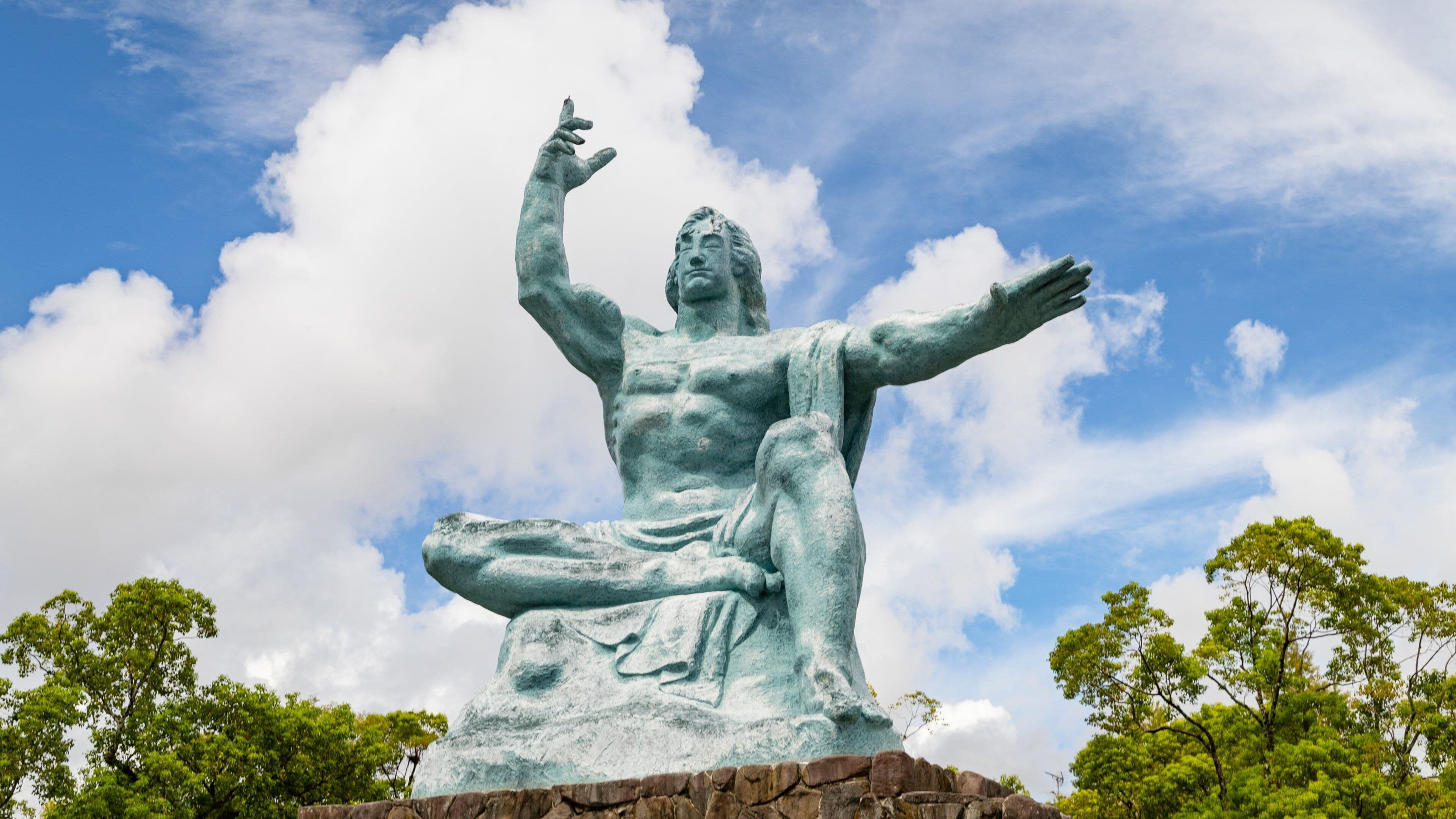 A large bronze statue of a seated figure with arms raised, set against a backdrop of a blue sky with clouds and green trees.
