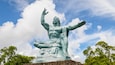 A large bronze statue of a seated figure with arms raised, set against a backdrop of a blue sky with clouds and green trees.