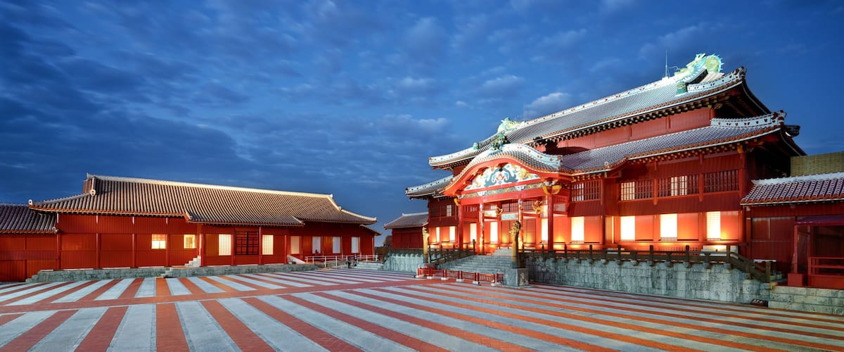 Un templo japonés tradicional con techo rojo y decoraciones elaboradas, un patio empedrado en piedra y un cielo azul despejado.