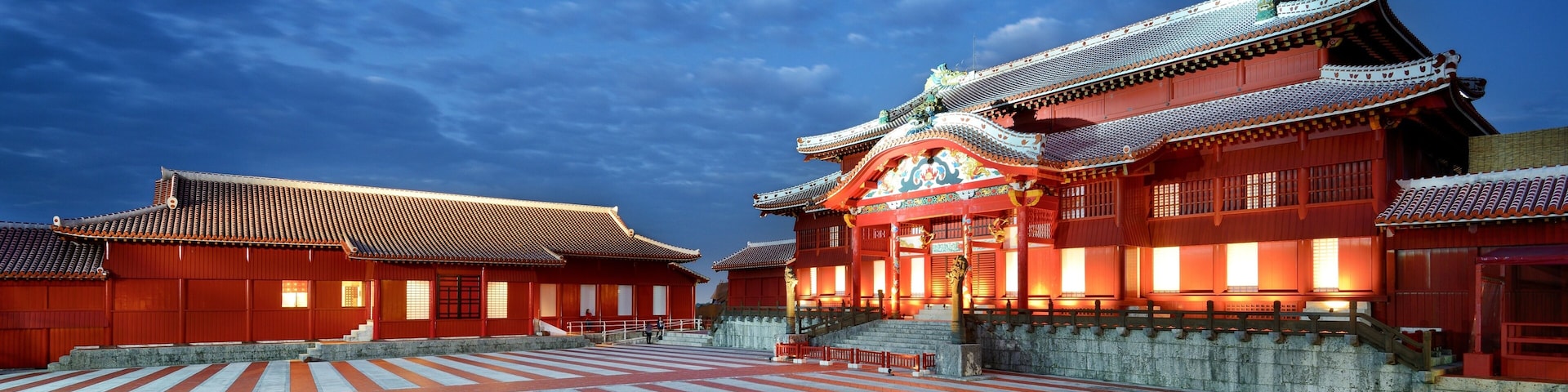 Um templo japonês tradicional com telhado vermelho e decorações elaboradas, pátio pavimentado em pedra e céu azul límpido.