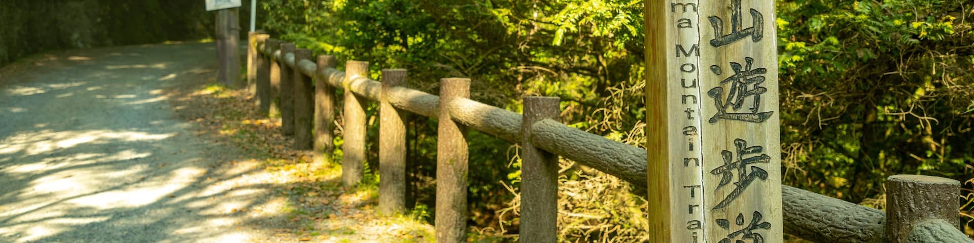 A wooden signpost with Japanese characters, a gravel path, and a wooden fence in a forested area.