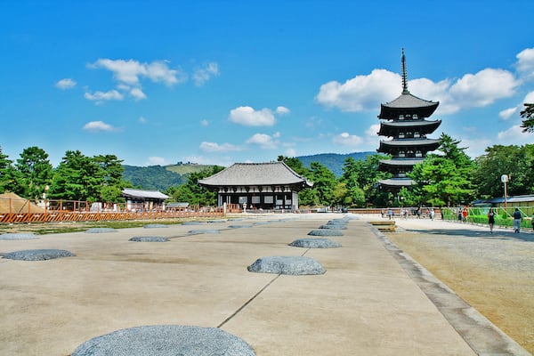 Ein traditioneller japanischer Tempel mit einer mehrstufigen Pagode, eingebettet in eine friedvolle Landschaft mit Bäumen und blauem Himmel.