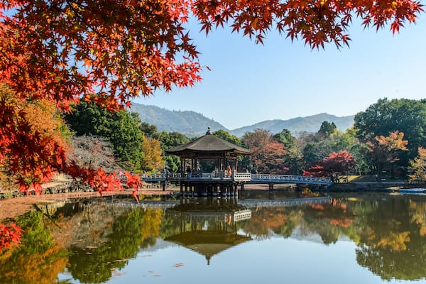 Ein traditionelles japanisches Pavillon über einem Teich, umgeben von Herbstlaub und Hügeln.