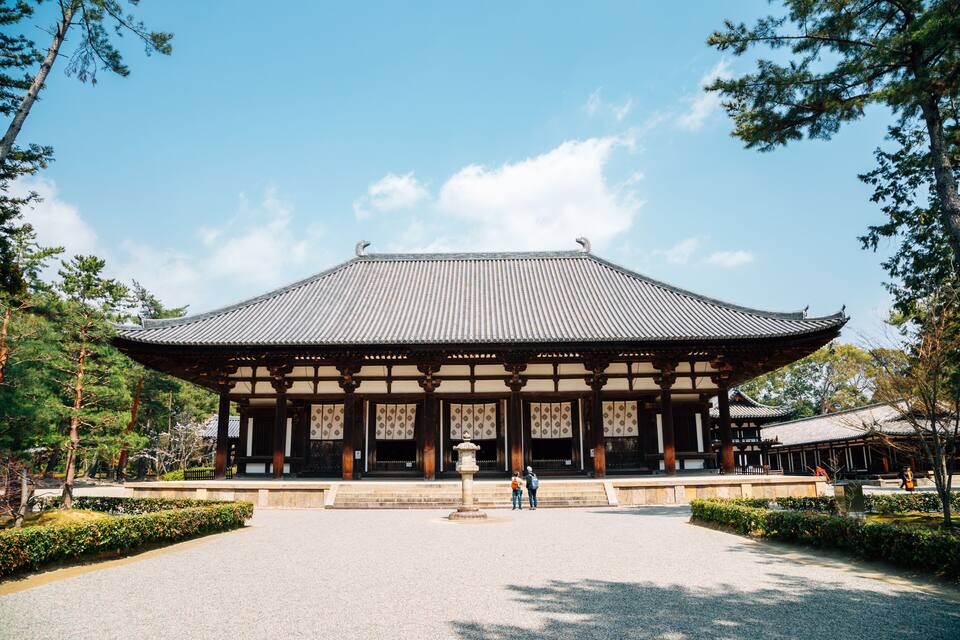 A traditional Japanese building with a tiled roof and wooden pillars, surrounded by a gravel courtyard and trees.