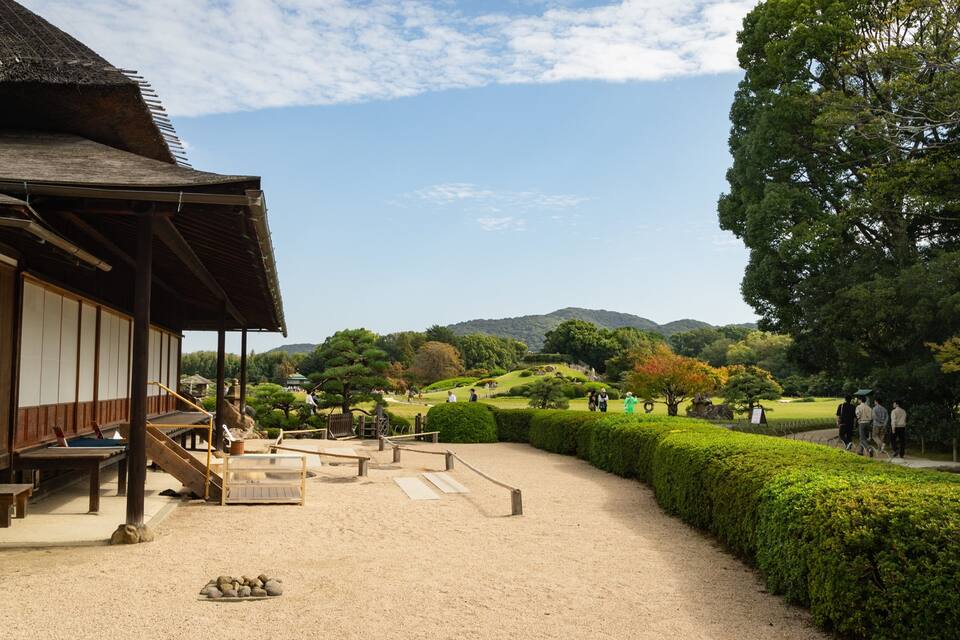Ein traditionelles japanisches Gebäude mit einem strohgedeckten Dach, einem Kiesweg, ordentlich geschnittenen Büschen und einer Bergkulisse.