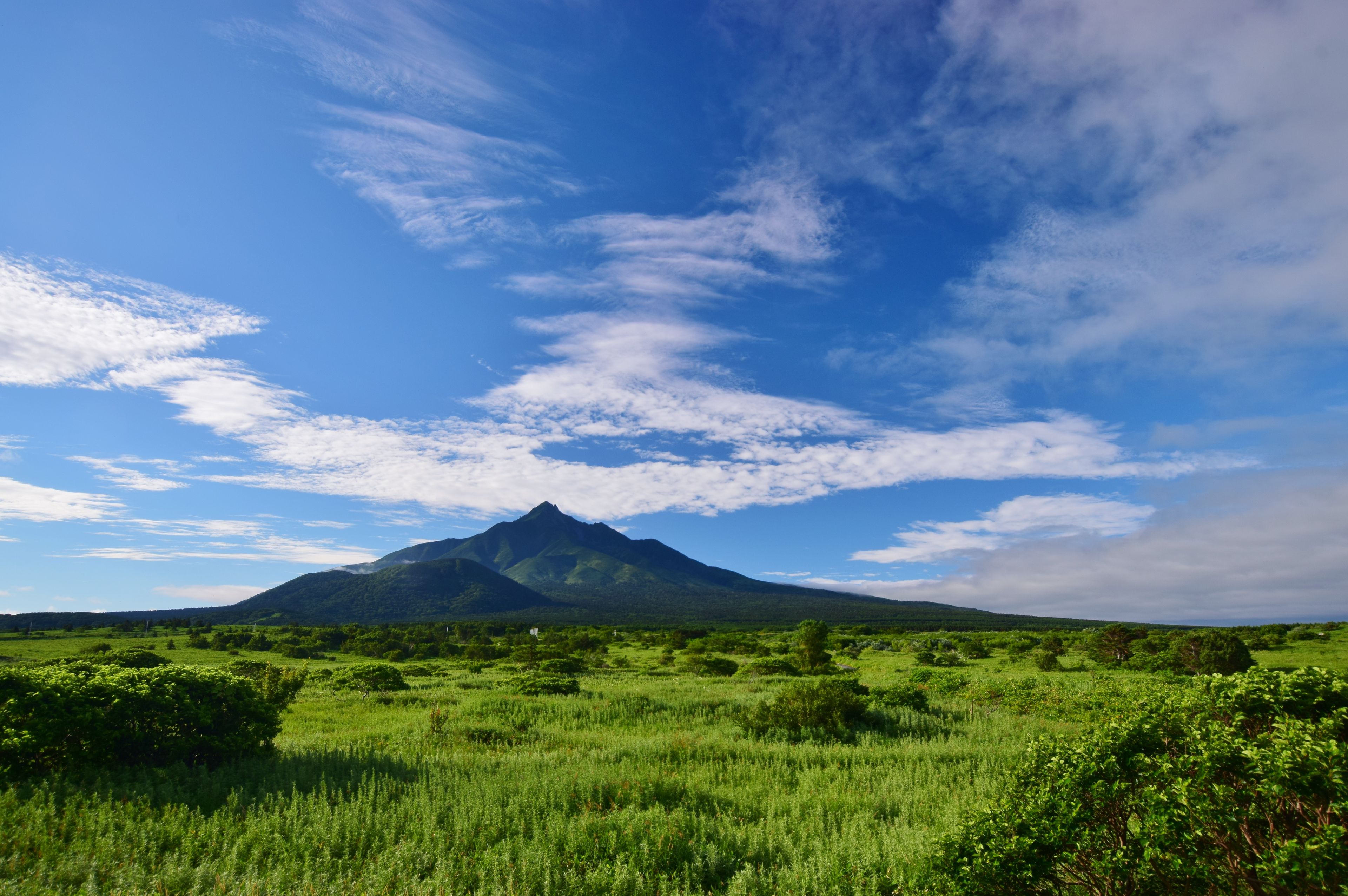 A mountain landscape with lush green fields and a clear blue sky with scattered clouds.