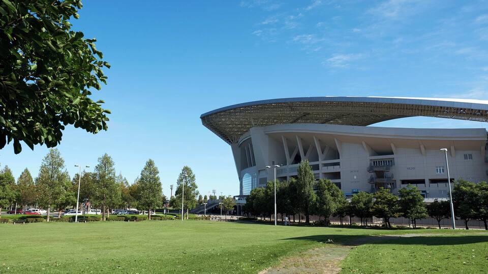 A modern stadium with a unique roof structure, surrounded by a well-maintained grassy area and trees.