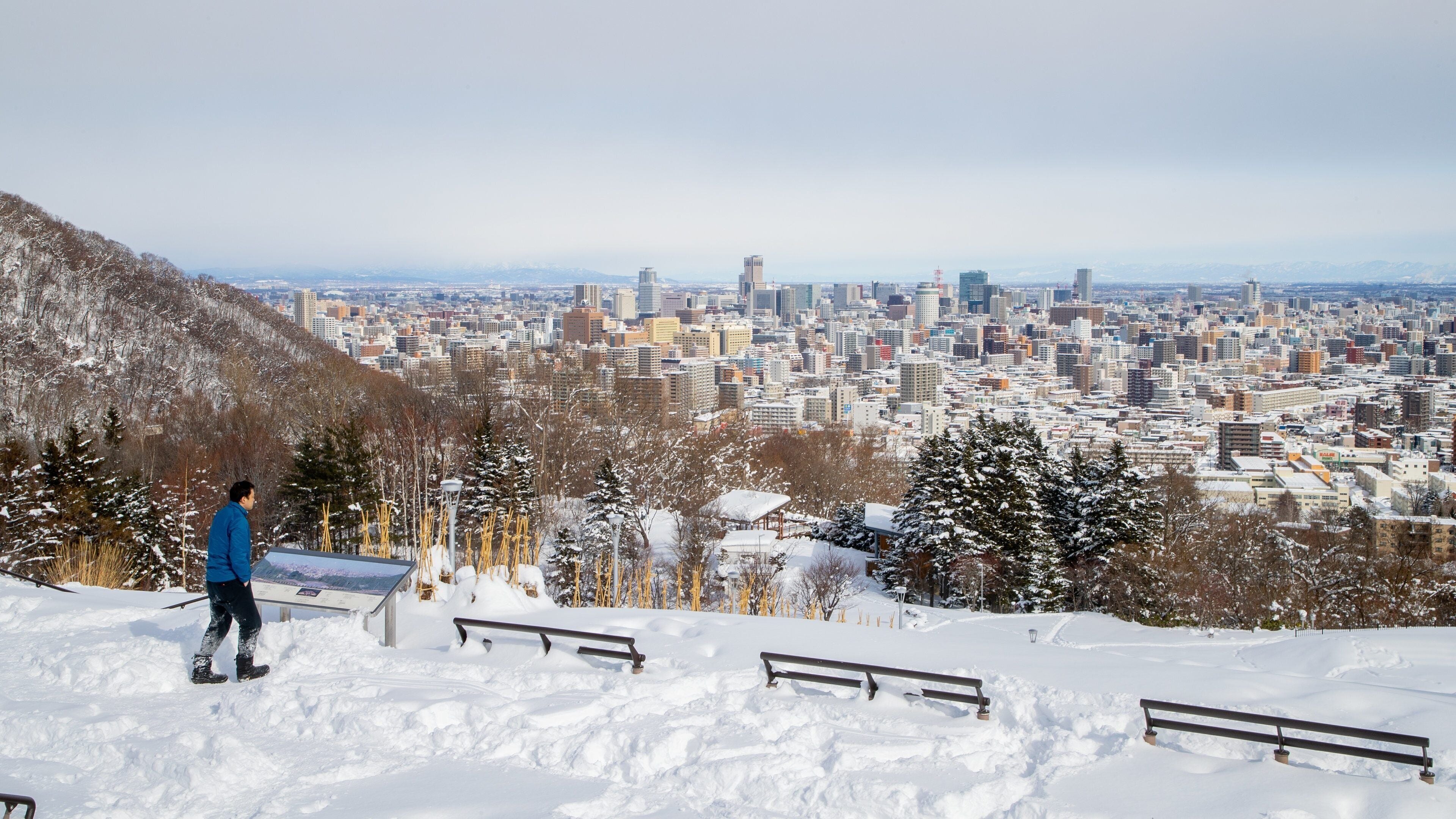 A snowy cityscape with a person walking on a snow-covered slope, benches, and a distant view of the city.
