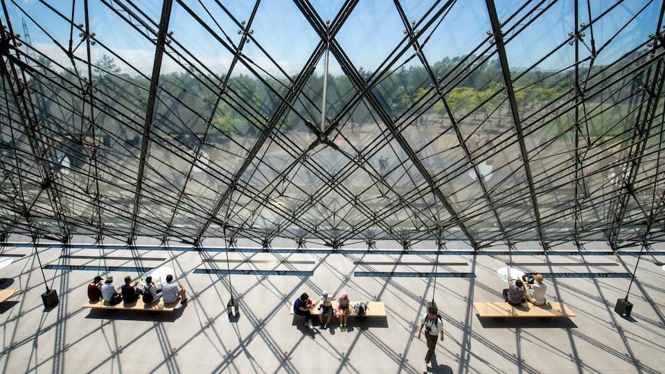 Una gran estructura de paneles de vidrio con un diseño geométrico, bancos y personas sentadas y caminando.