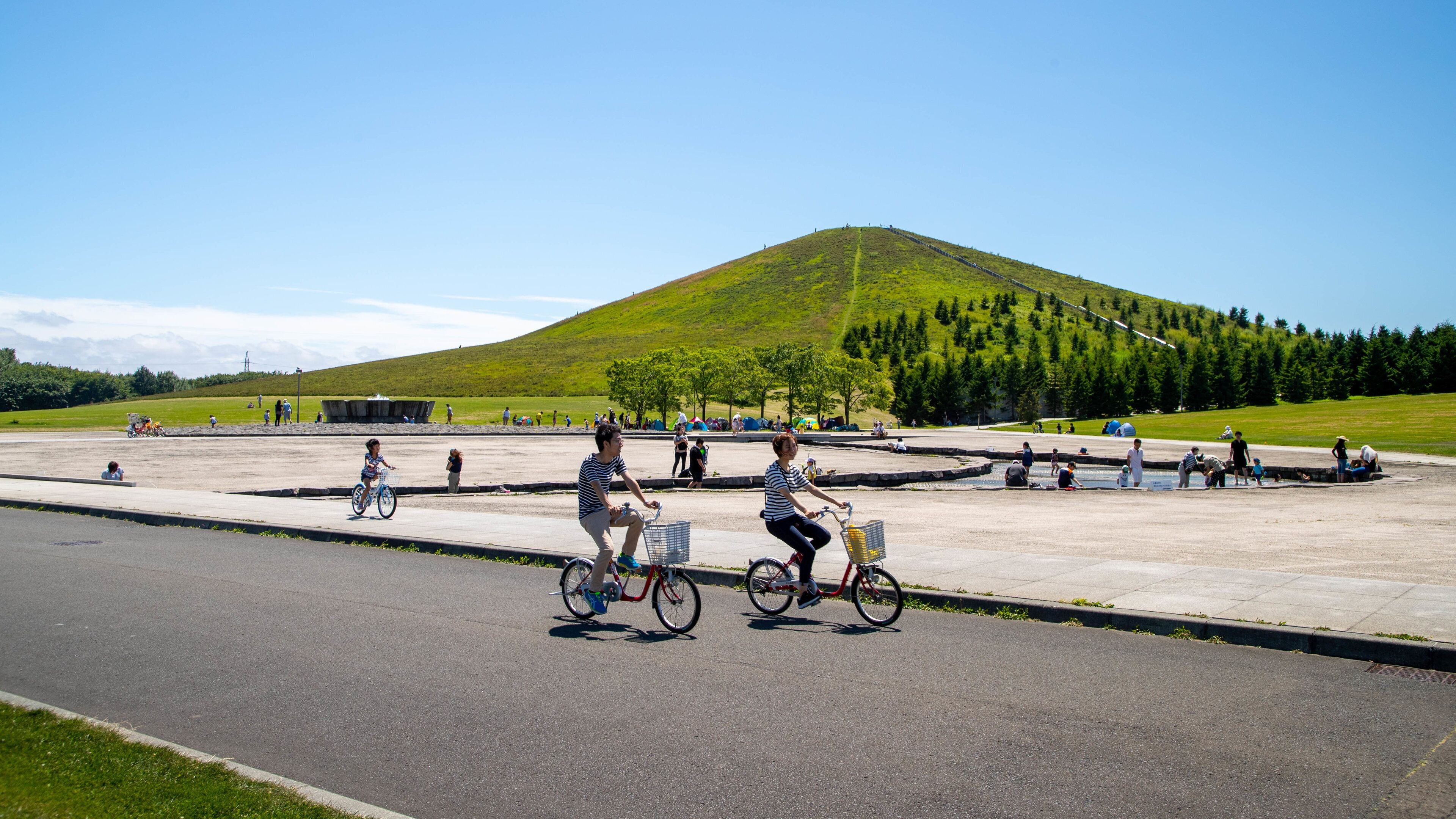 A paved area with people riding bicycles, a grassy hill, and trees in the background.