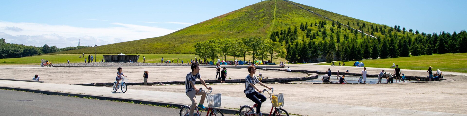A paved area with people riding bicycles, a grassy hill, and trees in the background.