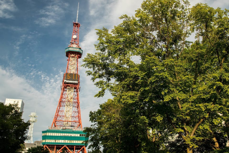 A tall red tower with a clock and the word "Panasonic" on it, surrounded by green trees.