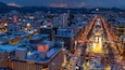 Un paisaje urbano al anochecer con edificios iluminados, una noria destacada y montañas nevadas en el fondo.