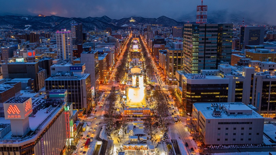 A cityscape with a prominent illuminated square at the center, surrounded by tall buildings and a snowy mountain backdrop.