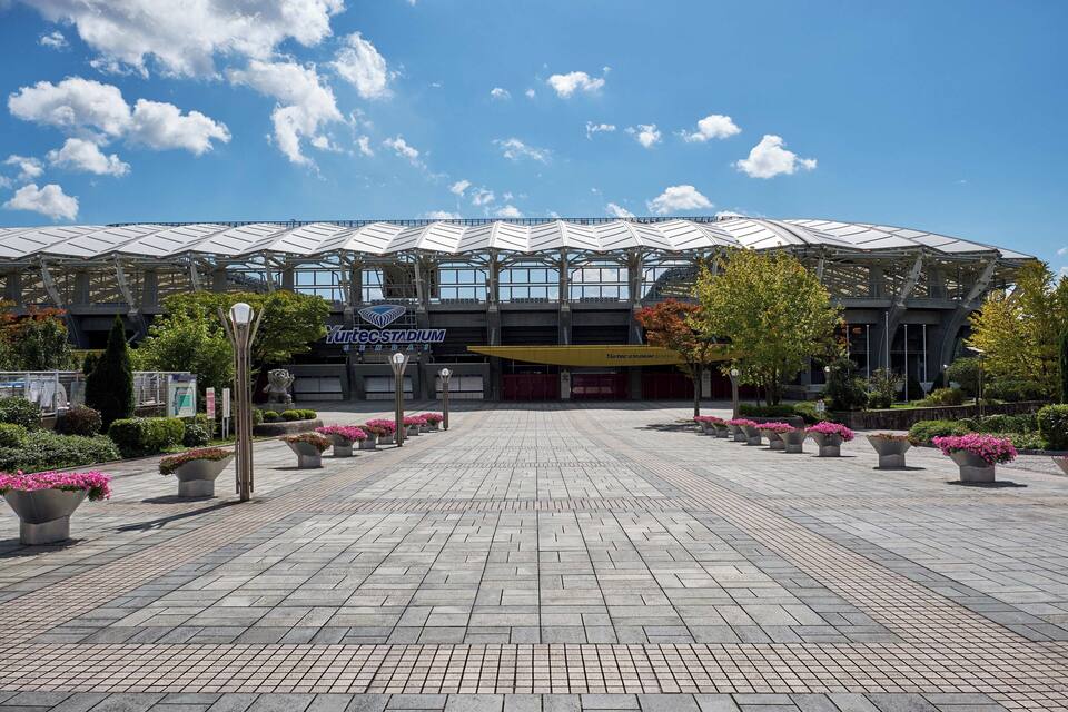 A stadium with a modern design, surrounded by potted plants and a clear blue sky.