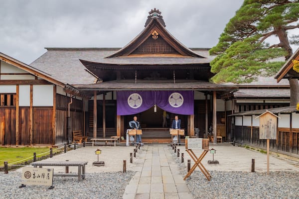 Ein traditionelles japanisches Gebäude mit einem Ziegeldach, Holzbalken und einem zentralen Eingang, der von zwei Holztüren gesäumt wird.