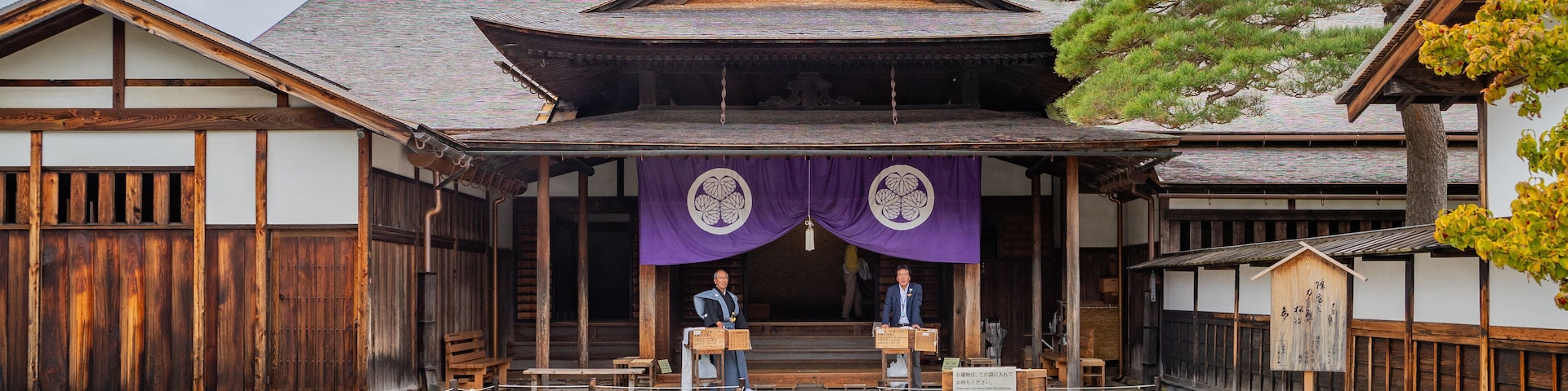 Ein traditionelles japanisches Gebäude mit einem Ziegeldach, Holzbalken und einem zentralen Eingang, der von zwei Holztüren gesäumt wird.