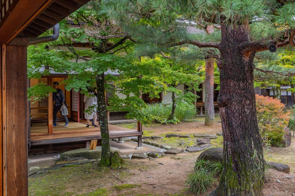 A traditional Japanese garden with a wooden building, a large tree, and a stone path.