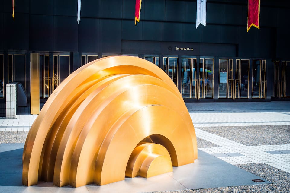 A modern architectural structure with a series of curved, golden panels in front of a building labeled 'Suntory Hall'.