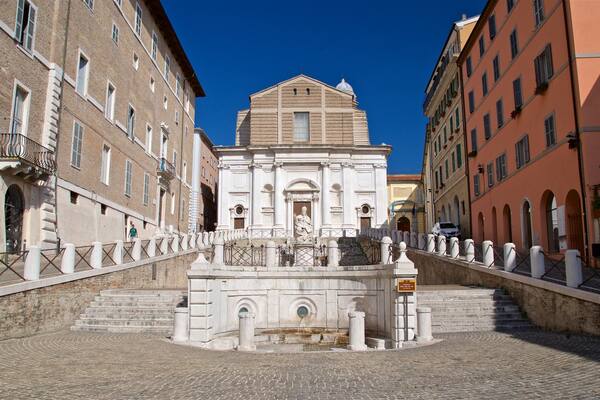 Une place historique, ornée d’une fontaine centrale, bordée de bâtiments de style classique et surplombée d’un ciel d’un bleu éclatant.