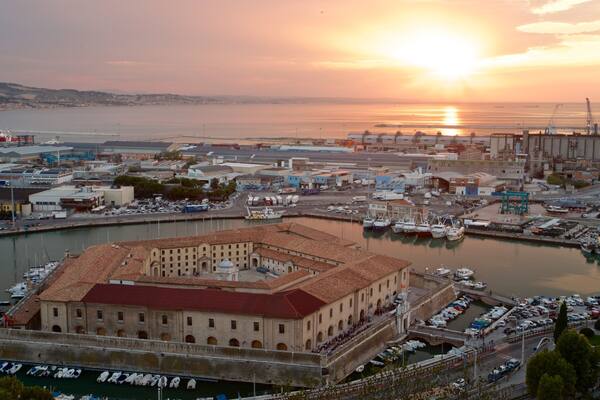 Un paysage urbain côtier comprenant un bâtiment historique remarquable, une marina avec des bateaux et des structures industrielles.