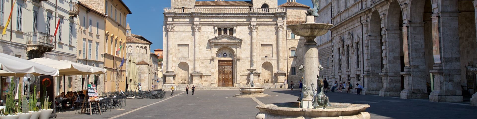 Una plaza histórica con una iglesia destacada que incluye un campanario, una fuente y edificios circundantes con banderas.