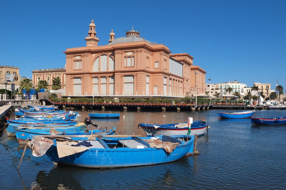 A harbor with several blue boats docked in front of a large pink building with domes.