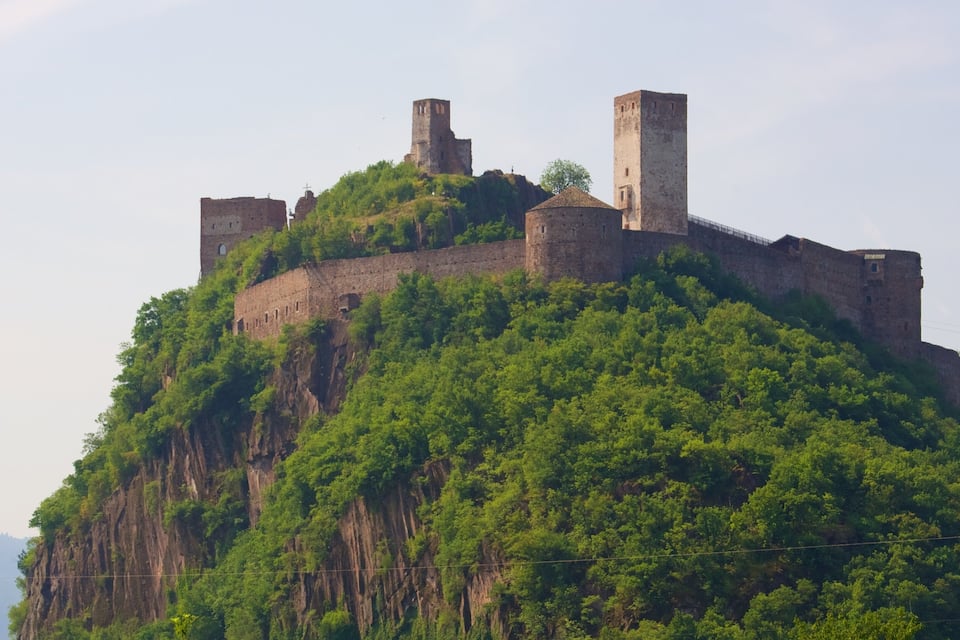 Un castell medieval al cim d'un turó verd i exuberant, amb diverses torres.