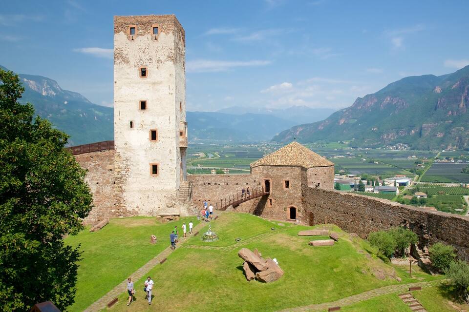 Un castell històric amb una torre alta, envoltat de vegetació exuberant i muntanyes al fons.