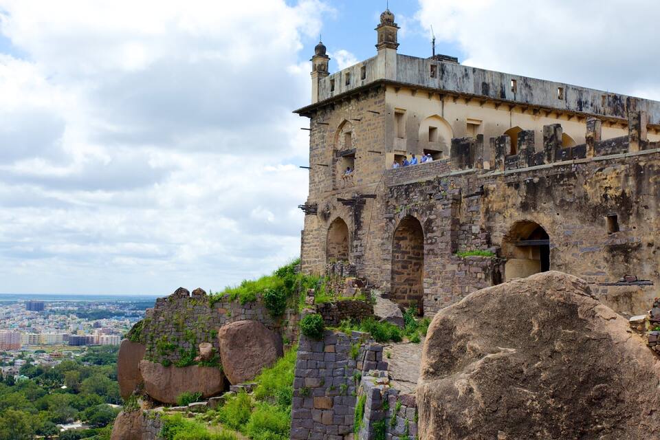 A historic stone fortress with arched windows and a tower, overlooking a cityscape.