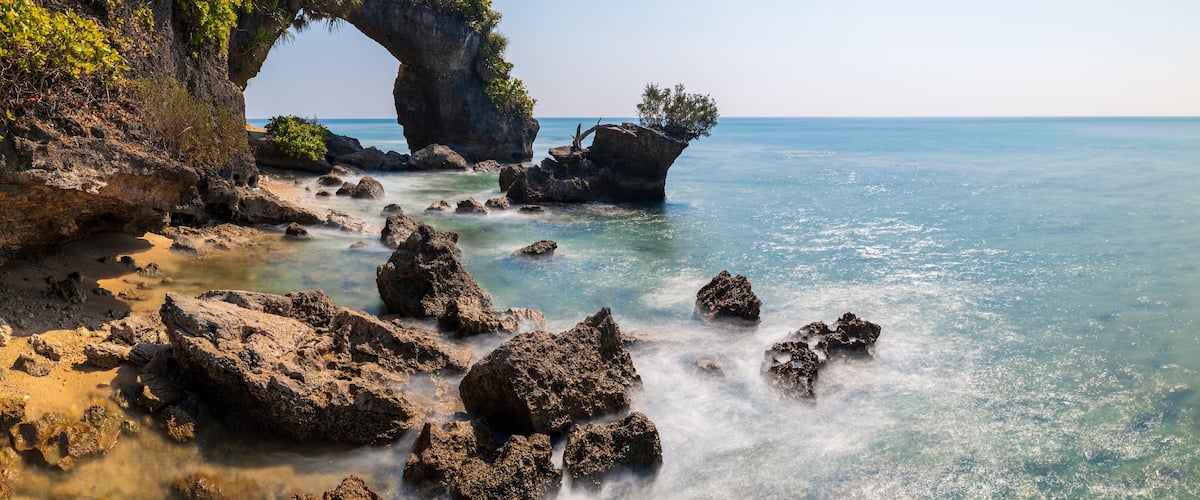 A coastal landscape with a prominent rock arch formation, clear blue water, and lush greenery.