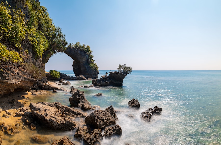 A coastal landscape with a prominent rock arch formation, clear blue water, and lush greenery.