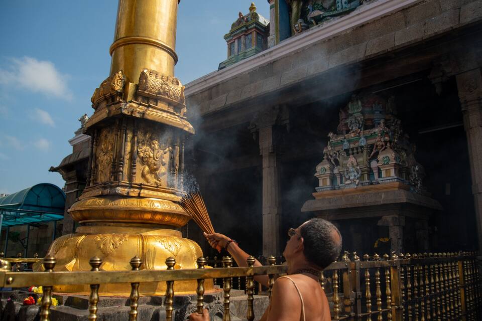 A golden statue with intricate carvings, a person performing a ritual, and traditional temple architecture in the background.