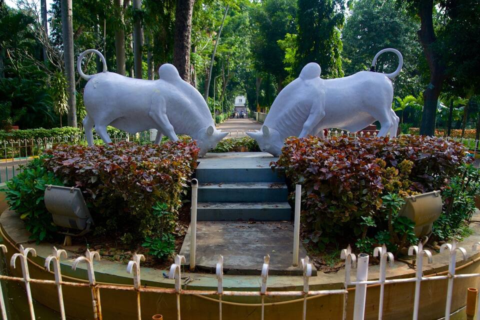 A park with two large white sculptures of bulls facing each other, surrounded by greenery and a fountain.