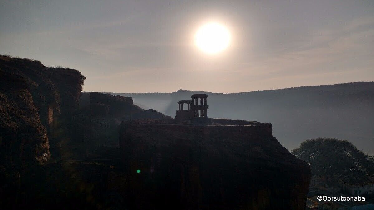 A historical structure silhouetted against the sun, with a mountainous backdrop and a clear sky.