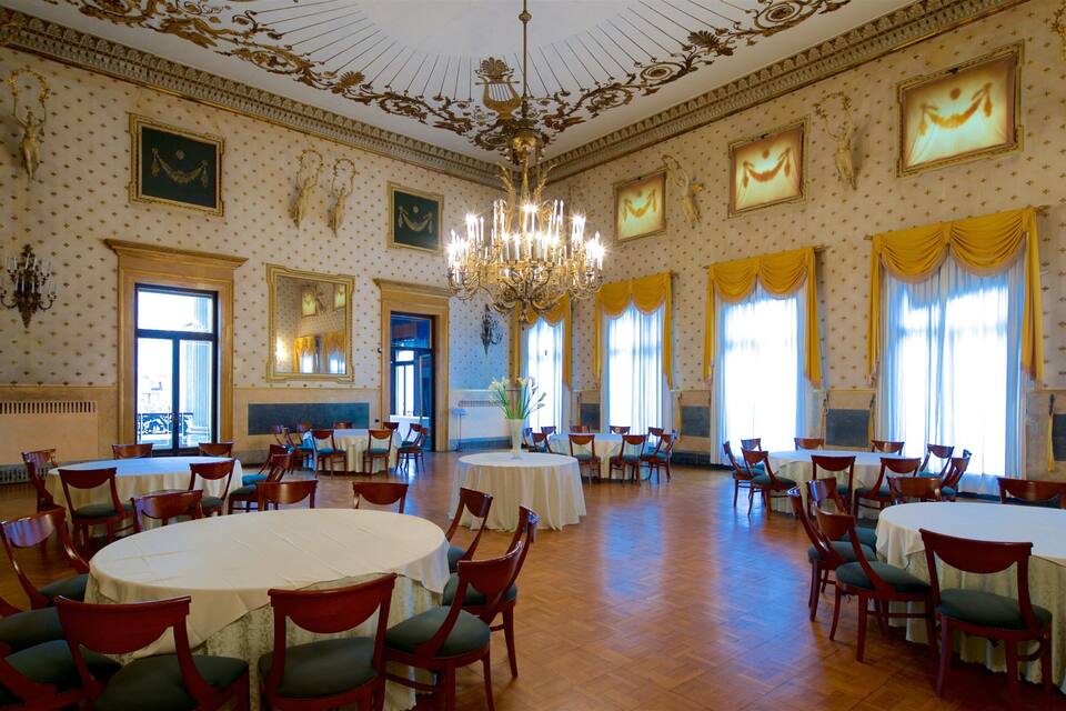 A grand hall with ornate chandeliers, large windows with draped curtains, and multiple round tables set for dining.