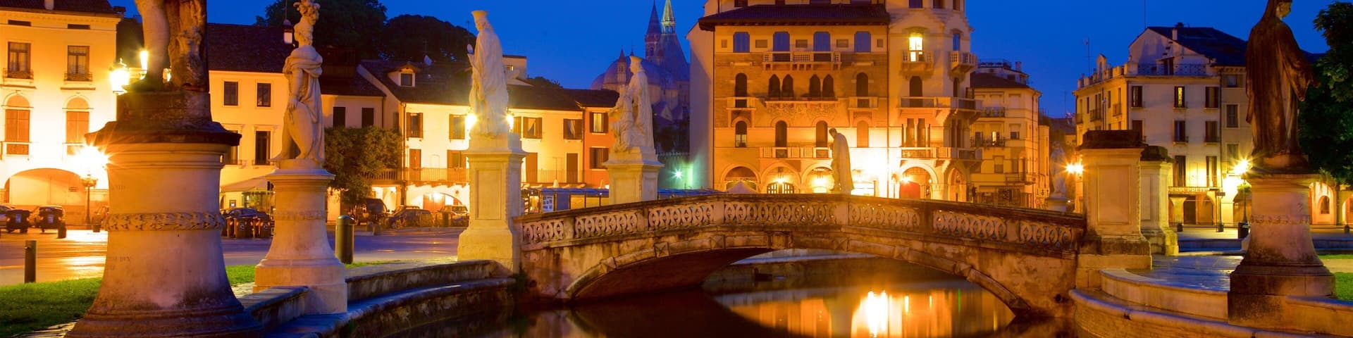 A historic square with a stone bridge over a canal, surrounded by classical statues and illuminated buildings.