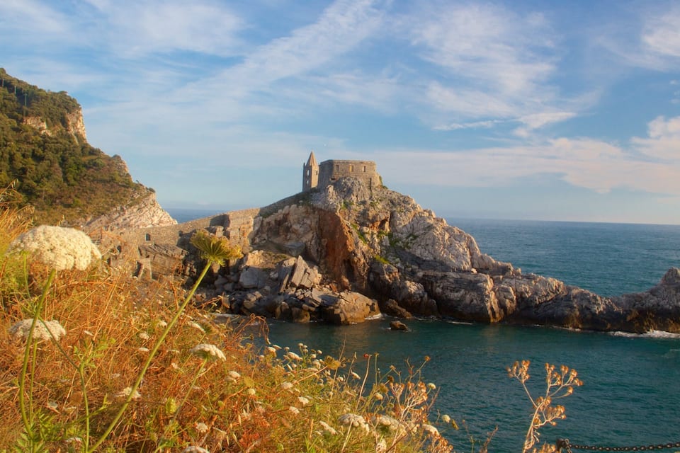A coastal landscape with a historic stone fortress on a rocky outcrop, surrounded by lush greenery and the sea.