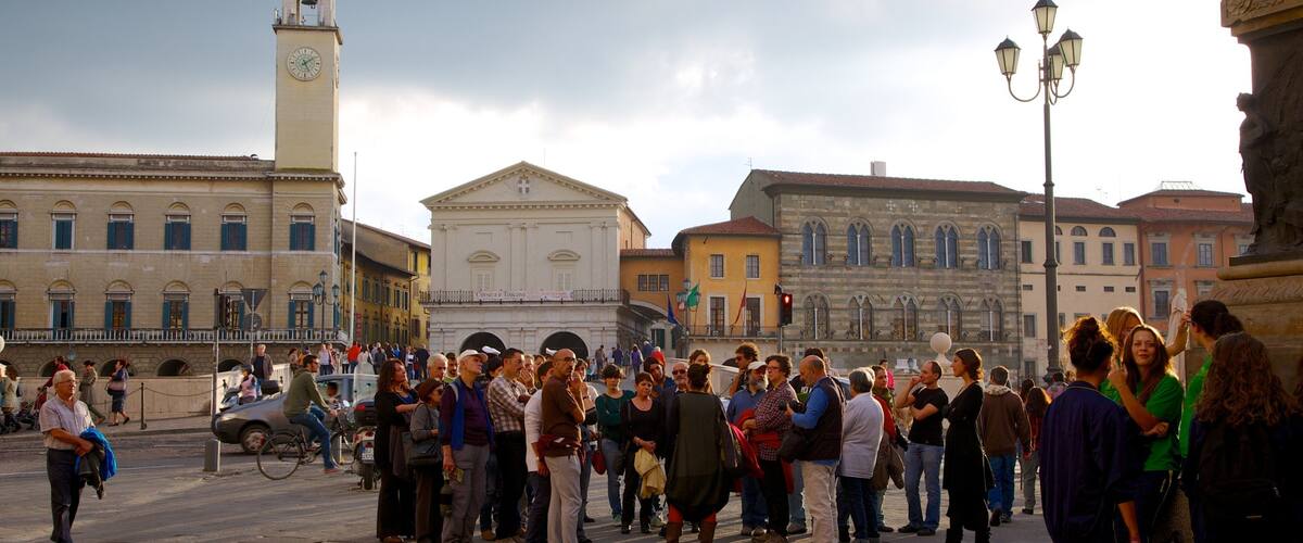 A crowded square with historic buildings, a clock tower, and a fountain.