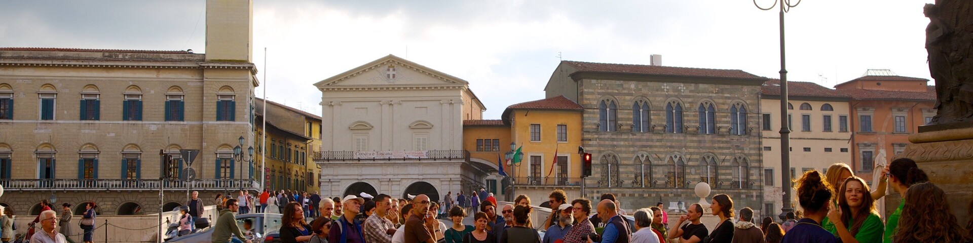 A crowded square with historic buildings, a clock tower, and a fountain.