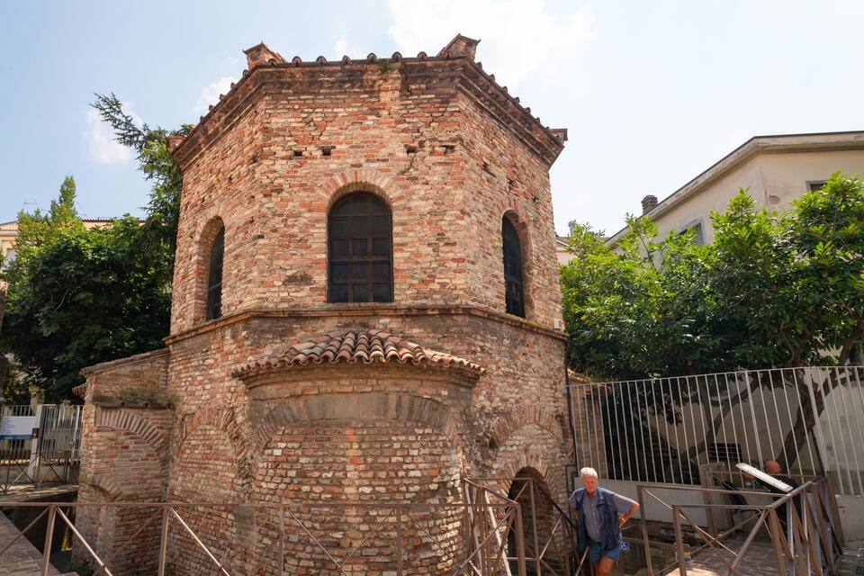 A historic brick tower with arched windows and a tiled roof, surrounded by trees and a metal fence.