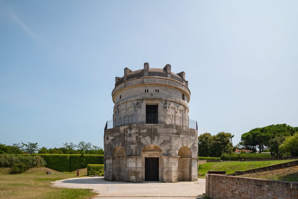 Ein historischer Stein-Turm mit kreisförmigem Fundament und einem Balkon, umgeben von Grünpflanzen und blauem Himmel.