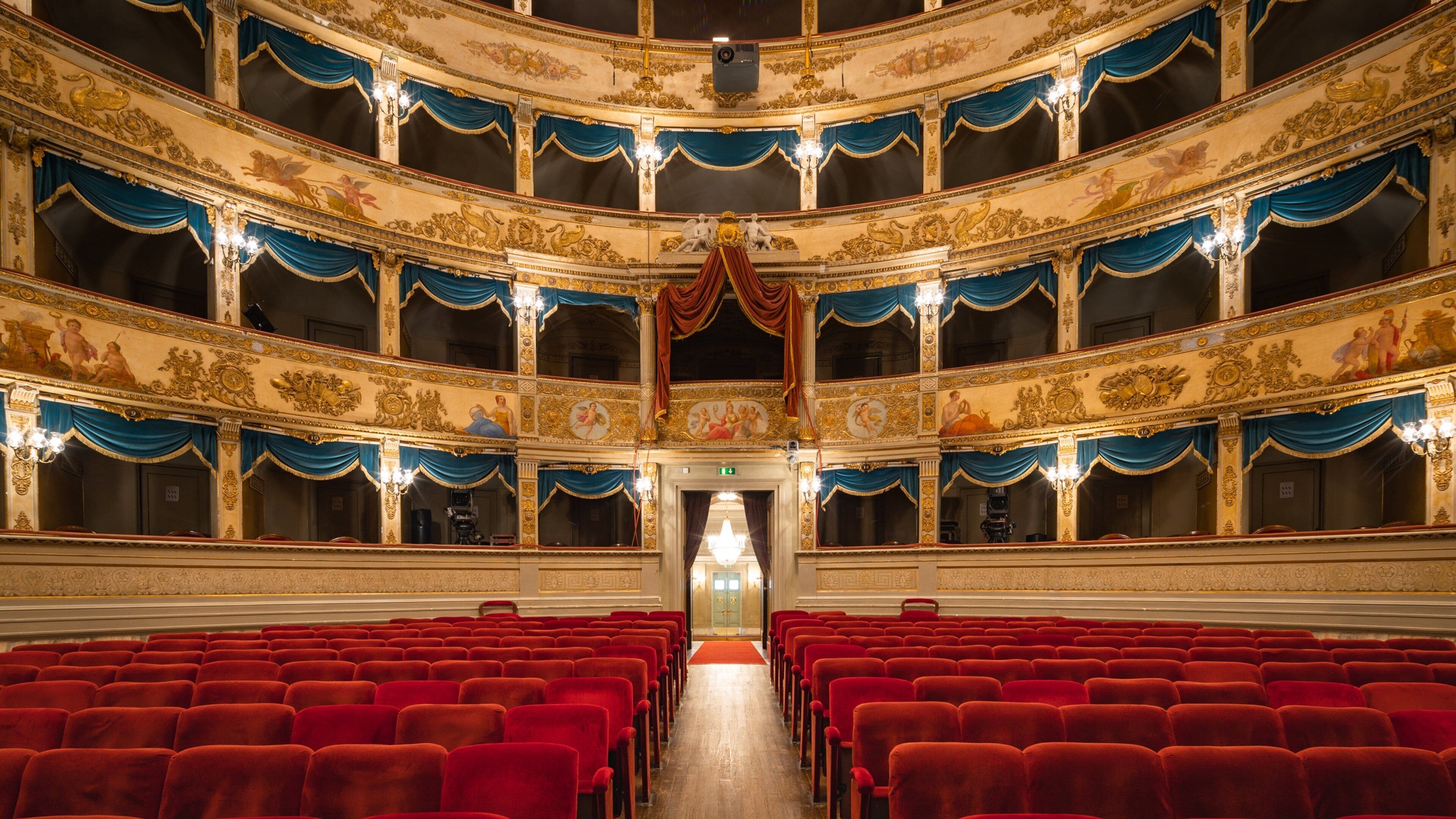 A grand theater with ornate gold and blue decorations, red velvet seats, and a central stage.